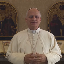 A man in religious attire speaks, standing in front of ornate book stands with open texts. Decorative gold and black backgrounds.