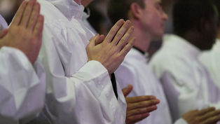 People in white robes stand with hands clasped in prayer. The setting is a solemn indoor gathering with soft lighting, creating a serene mood.