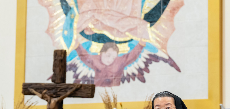 Nun speaking at a lectern in a church, with a crucifix, flowers, and a large Virgin Mary mural behind her.