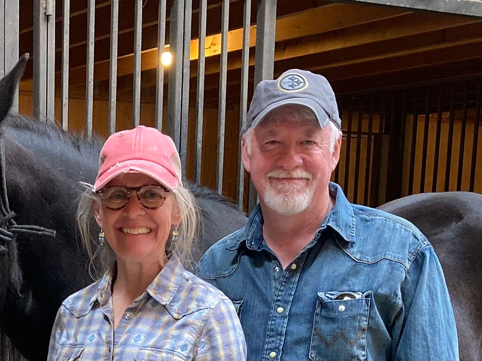 A man and woman smile in a barn, wearing caps and denim shirts. A horse is partially visible beside them. Warm lighting and metal bars in the background.