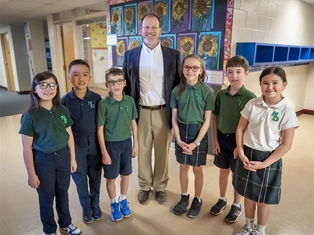 A teacher poses with six students in a school hallway. They wear green and white uniforms, standing in front of colorful sunflower artwork, smiling.