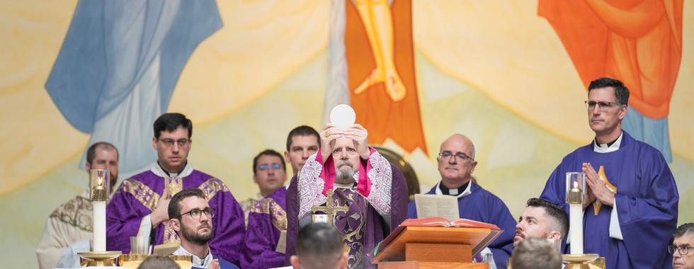 Bishop holds up a communion wafer during mass, surrounded by clergy in purple vestments. A mural with religious imagery is in the background.