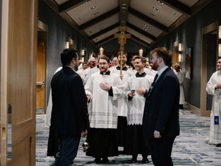Catholic seminarians in white vestments walk in procession down a hallway, carrying candles and a crucifix, as attendees stand on either side greeting them.