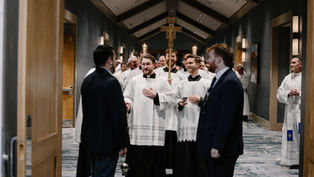 Catholic seminarians in white vestments walk in procession down a hallway, carrying candles and a crucifix, as attendees stand on either side greeting them.