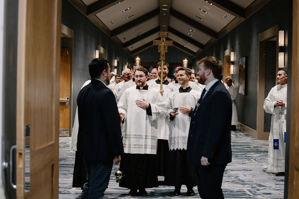 Catholic seminarians in white vestments walk in procession down a hallway, carrying candles and a crucifix, as attendees stand on either side greeting them.