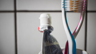 Toothpaste tube and two colorful toothbrushes in a cup against a tiled bathroom wall.
