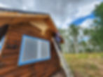 Person on a ladder staining a wooden cabin in a forest. Sky is cloudy with patches of blue. Cabin windows covered for protection.