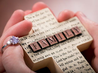 Hands holding a beige cross embossed with text and "FAMILY" in brown blocks. A ring with a sparkling stone is on one finger.