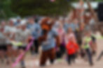 Runners, including a person in a buffalo costume, race outdoors on a dirt path, surrounded by spectators. Energetic atmosphere, colorful attire.