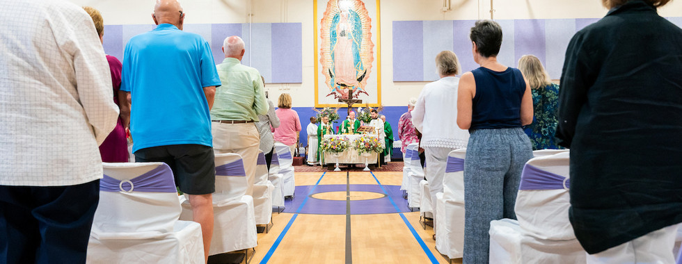 People stand for Mass in a gymnasium, facing priests at a flower-covered altar beneath a large Virgin Mary banner.
