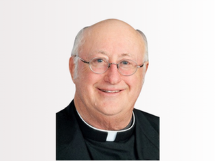 Smiling bald man in clergy attire with glasses, against a white background. He wears a black robe with a white clerical collar.