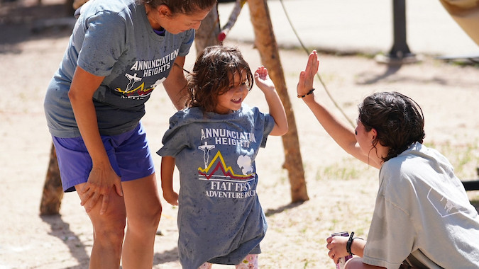 Three people outdoors, one adult bending, another crouched giving a high-five to child in a "Fiat Fest" shirt. Lively, sunny setting.