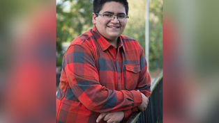 Smiling person in glasses and red plaid shirt leans on railing outdoors, with blurred greenery in the background, creating a relaxed mood.