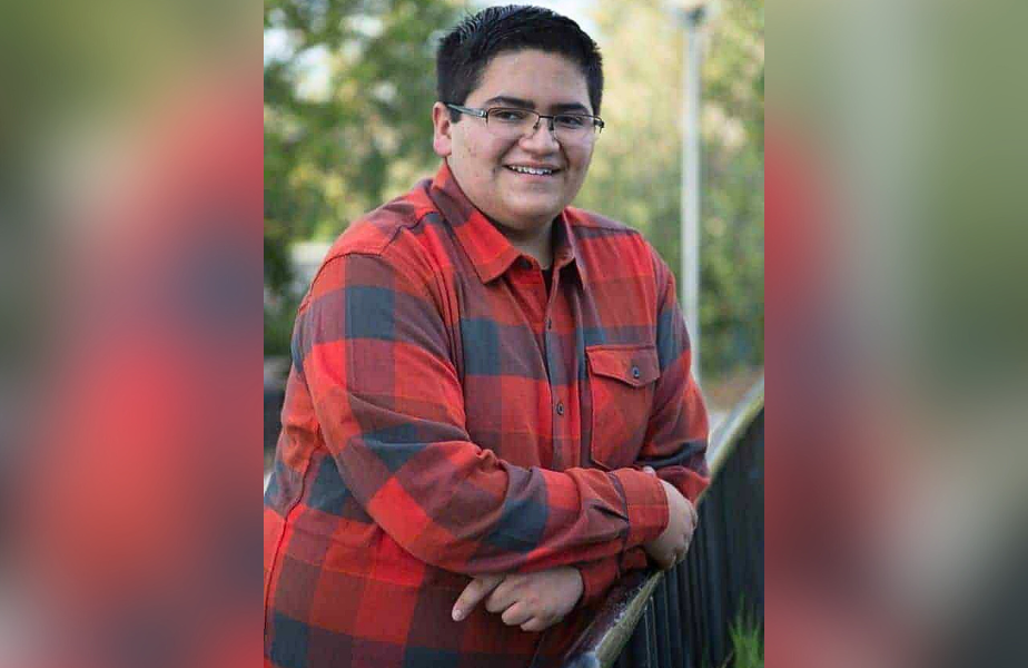 Smiling person in glasses and red plaid shirt leans on railing outdoors, with blurred greenery in the background, creating a relaxed mood.