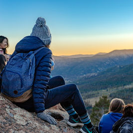 People sitting on a mountain ledge at sunset, wearing winter clothing. Blue sky, distant mountains, and a relaxed, contemplative mood.