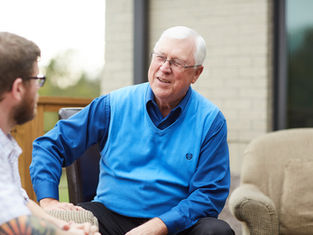 Elderly man in blue vest smiling while talking to another person outdoors, with a brick wall and porch furniture in the background.