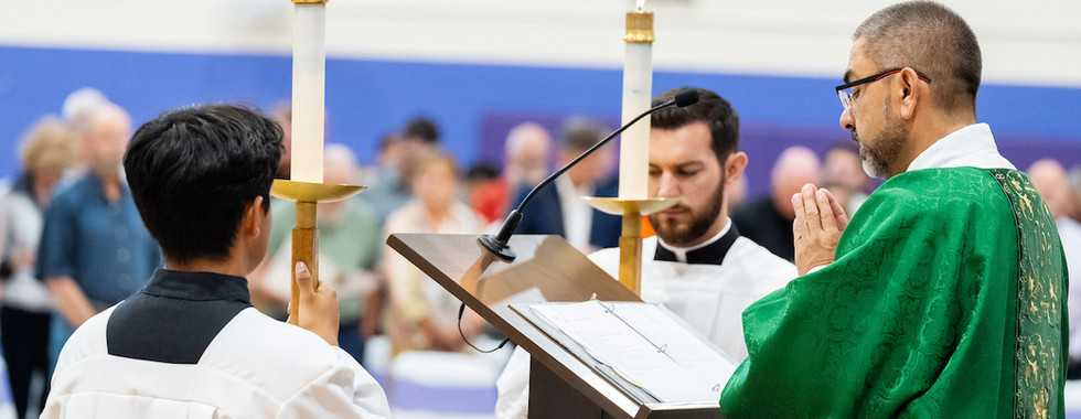 Deacon in green vestments prays at church lectern while two acolytes hold lit candles, with congregation blurred behind.
