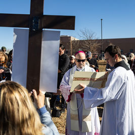 People in religious attire gather outside around a large cross on a sunny day. One person reads from a book, while others observe solemnly.