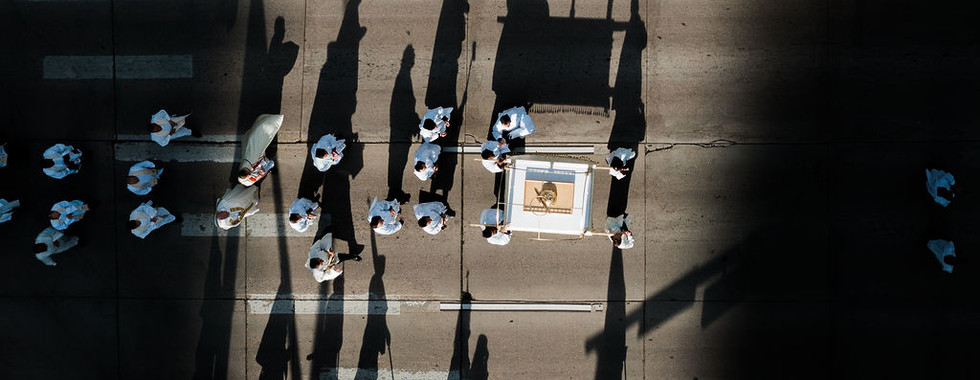 Aerial view of marchers in white on a city street, casting long shadows across the pavement in bright sunlight.