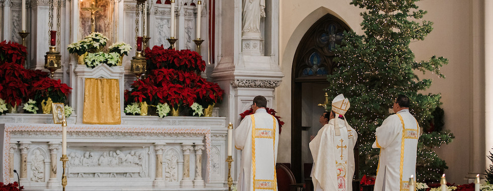 Clergy in white vestments stand before an ornate altar with poinsettias and a Christmas tree. Sacred and solemn church scene.
