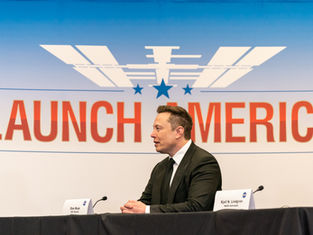 Man in a suit sits at a table with a "Launch America" backdrop. Two nameplates, one labeled Elon Musk and a NASA logo, are visible.