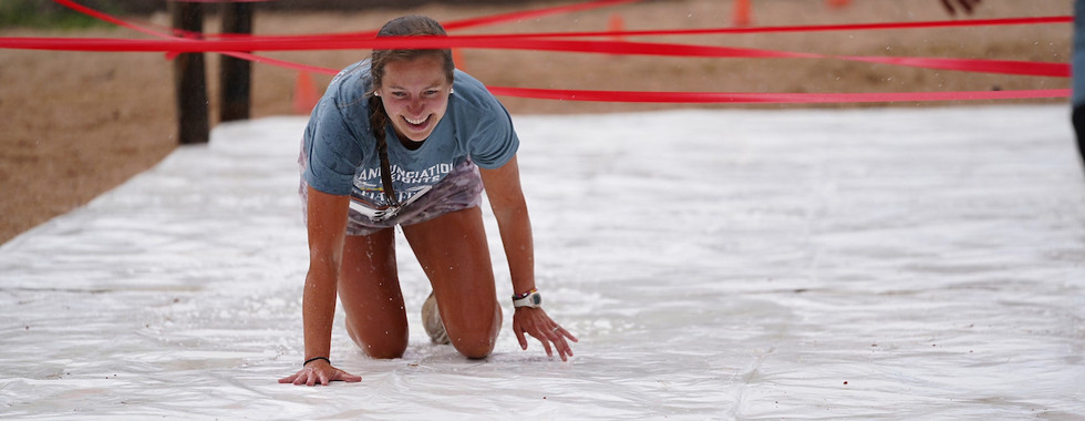 Smiling woman crawling under red ropes on a wet tarp, wearing a blue shirt. The setting appears outdoors with orange cones in the background.