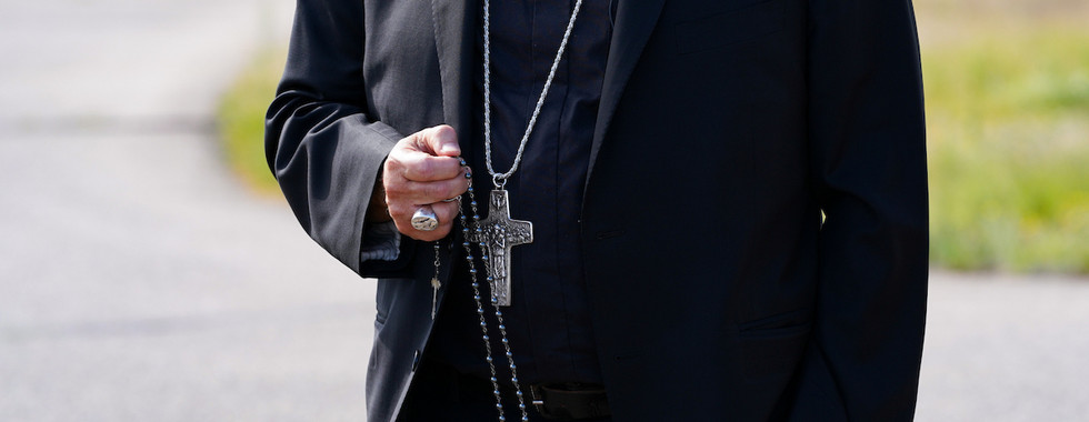 Clergyman in black suit holds a rosary with cross outdoors on a road, calm and solemn.