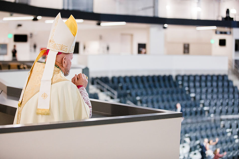 A religious figure in ornate robes and a mitre overlooks an empty auditorium from a balcony. Bright lights illuminate the modern space.
