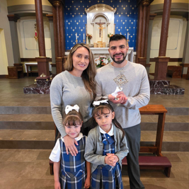 A family of four stands in a church. Parents in gray, daughters in plaid uniforms. The father holds a religious item. Blue altar backdrop.