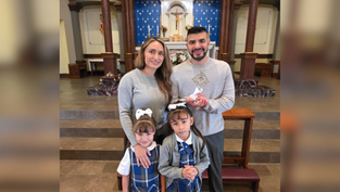A family of four stands in a church. Parents in gray, daughters in plaid uniforms. The father holds a religious item. Blue altar backdrop.