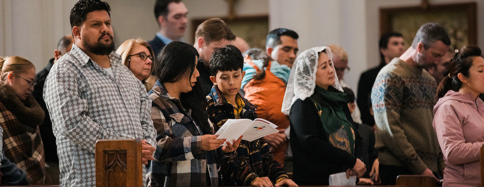 People standing in a church pew, holding papers, praying. Diverse attire, solemn mood. White walls, wooden pews, cross in background.