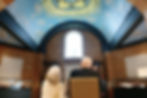 Two people sit in a room with a blue, decorative dome ceiling. They face a large arched window, surrounded by brick walls.
