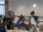 A nun speaks at a podium in a room with attendees seated at tables. Some guests take photos with phones. The mood is attentive.