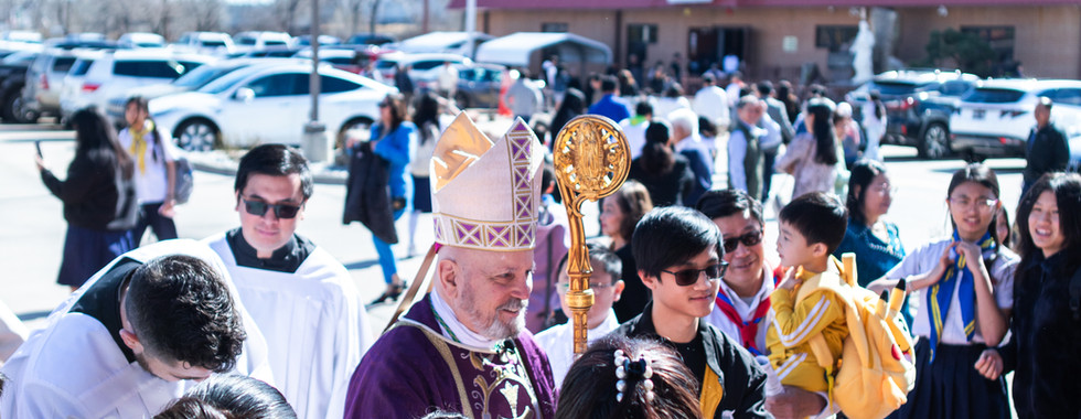 A religious procession with a bishop in purple robes and people gathered outside a building on a sunny day. Children and adults appear joyful.