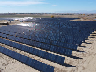 Rows of solar panels in a vast, open field under a clear blue sky. The panels reflect sunlight, with mountains visible in the distance.