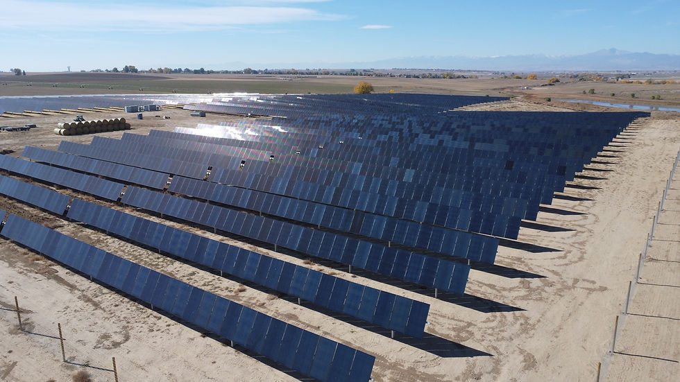 Rows of solar panels in a vast, open field under a clear blue sky. The panels reflect sunlight, with mountains visible in the distance.