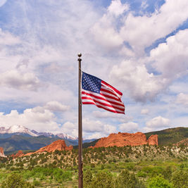 American flag on a pole above red rock mountains and green hills under a bright, cloudy blue sky.