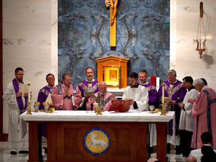 Priests in purple and pink vestments conduct a ceremony at an altar. A crucifix is on the bluish marble wall behind them.