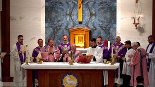 Priests in purple and pink vestments conduct a ceremony at an altar. A crucifix is on the bluish marble wall behind them.