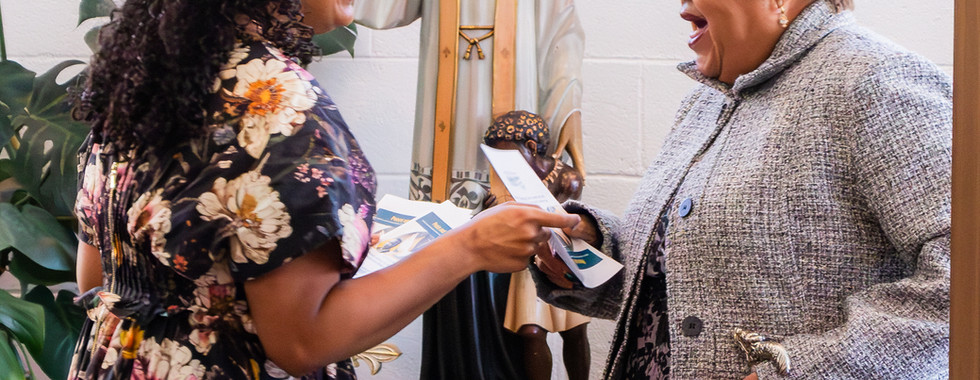 Two women smiling and exchanging papers in front of a religious statue. A sign reads "JESUS IS CALLING ALL YOUTH" on the wall.