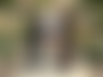 Two smiling men exchange a large check outdoors by a statue, surrounded by bushes and red flowers, with a building in the background.