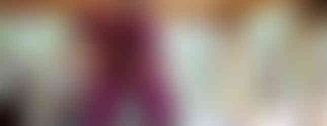 Man with mustache carrying girl on shoulders. Both wave and smile. Girl in pink outfit with face paint. Wooden structure and forest in background.