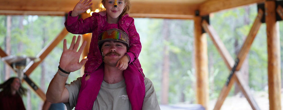 Man with mustache carrying girl on shoulders. Both wave and smile. Girl in pink outfit with face paint. Wooden structure and forest in background.
