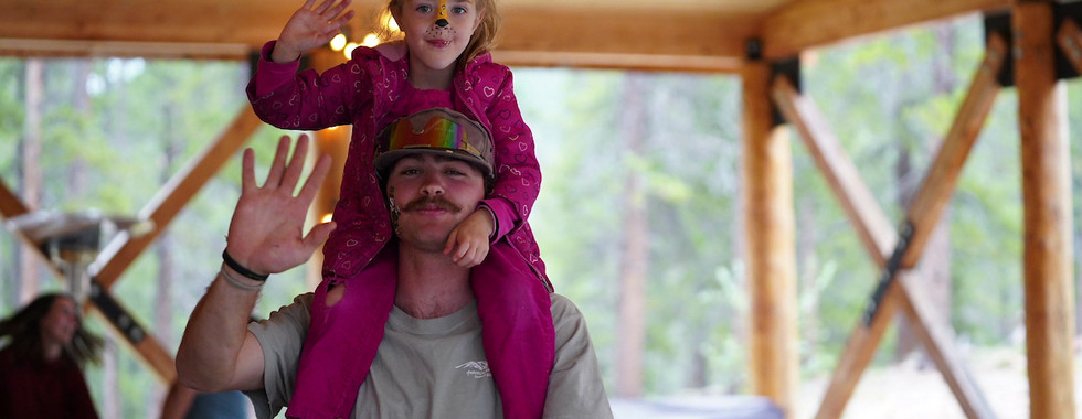 Man with mustache carrying girl on shoulders. Both wave and smile. Girl in pink outfit with face paint. Wooden structure and forest in background.