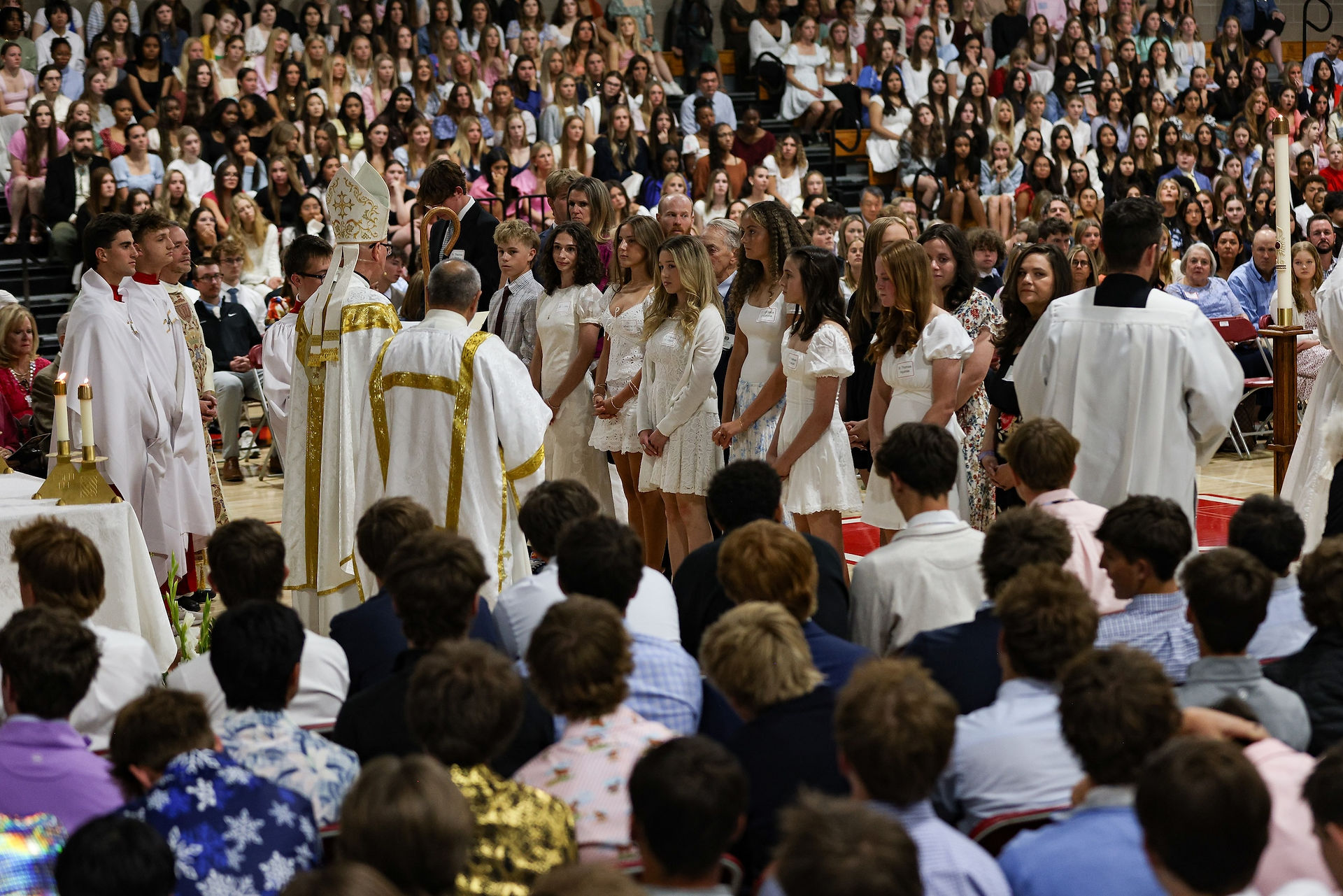 Catholic bishop blesses a large group of students gathered during a Catholic school Mass as hundreds of attendees watch from the bleachers.