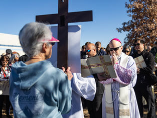 A priest in white robes and sunglasses prays before a large cross outdoors. A crowd, some with prayer books, stands solemnly under a clear sky.