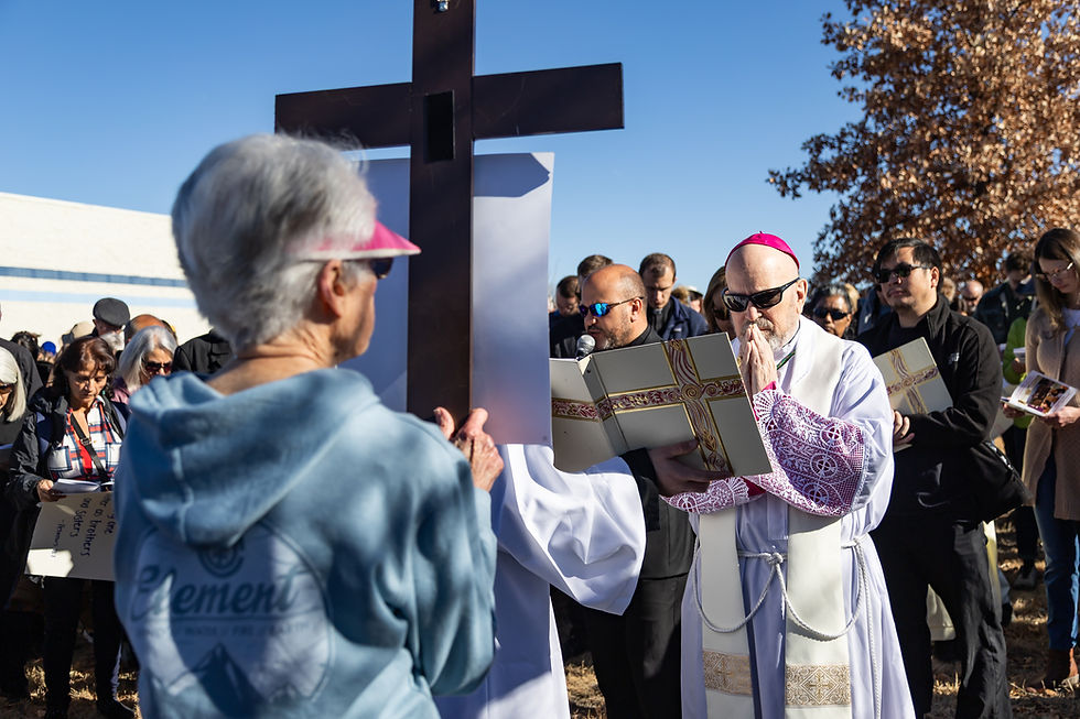 A priest in white robes and sunglasses prays before a large cross outdoors. A crowd, some with prayer books, stands solemnly under a clear sky.
