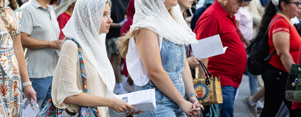 Crowd at an outdoor religious procession, with women in lace head coverings holding papers and walking solemnly.