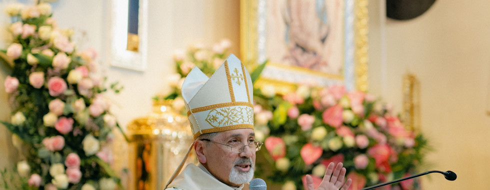 A bishop in white and gold vestments speaks at a church altar, surrounded by flowers, a crucifix, and religious art.