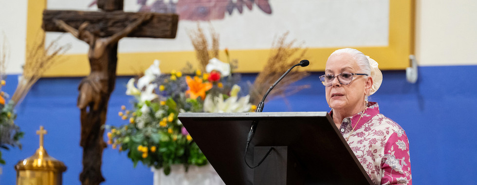 Elderly woman speaks at a lectern in a church, beside a crucifix and flowers, with a colorful angel mural in the background.